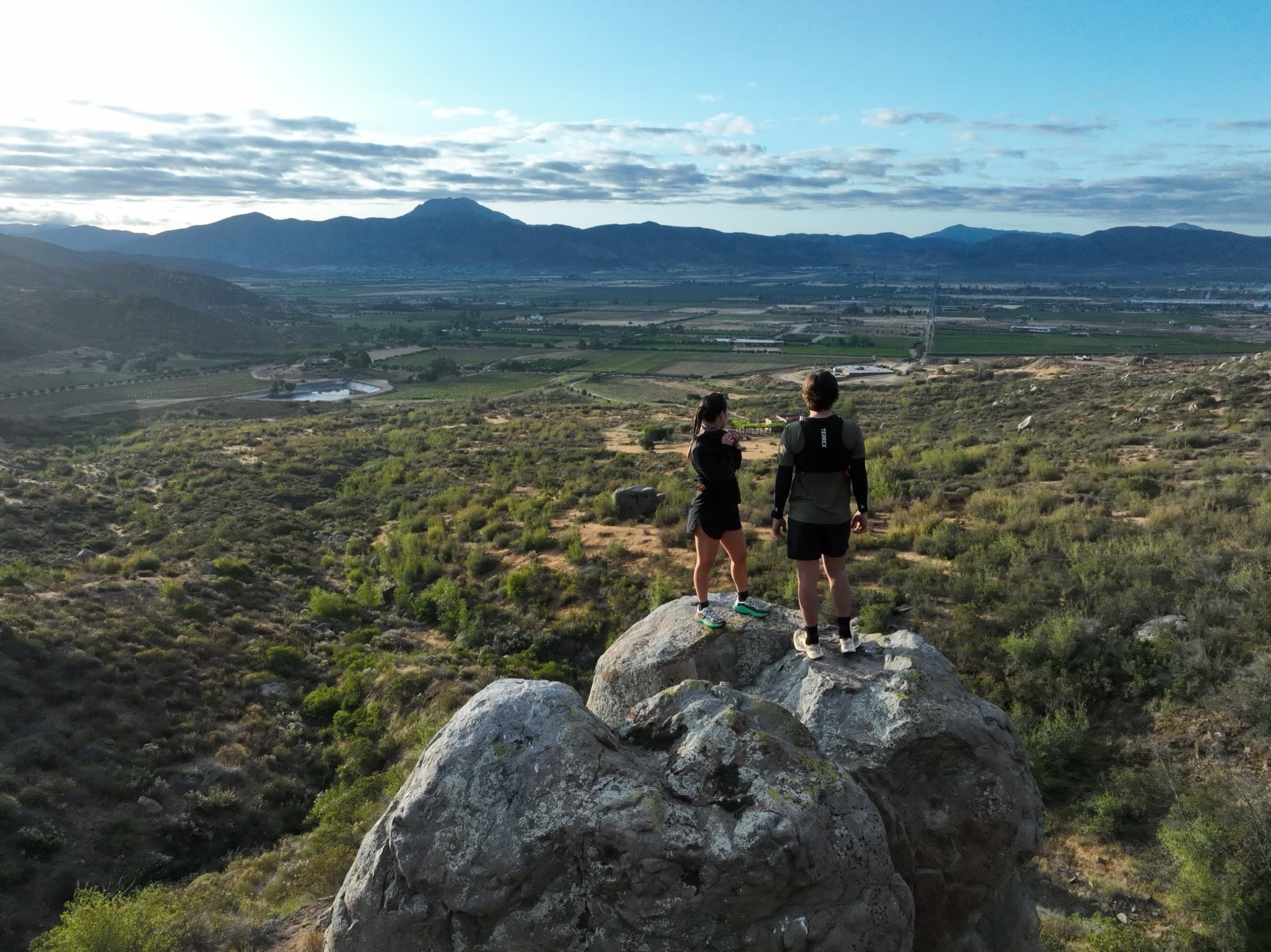Two runners look out over Valle de Guadalupe's vineyards from a rocky overlook.
